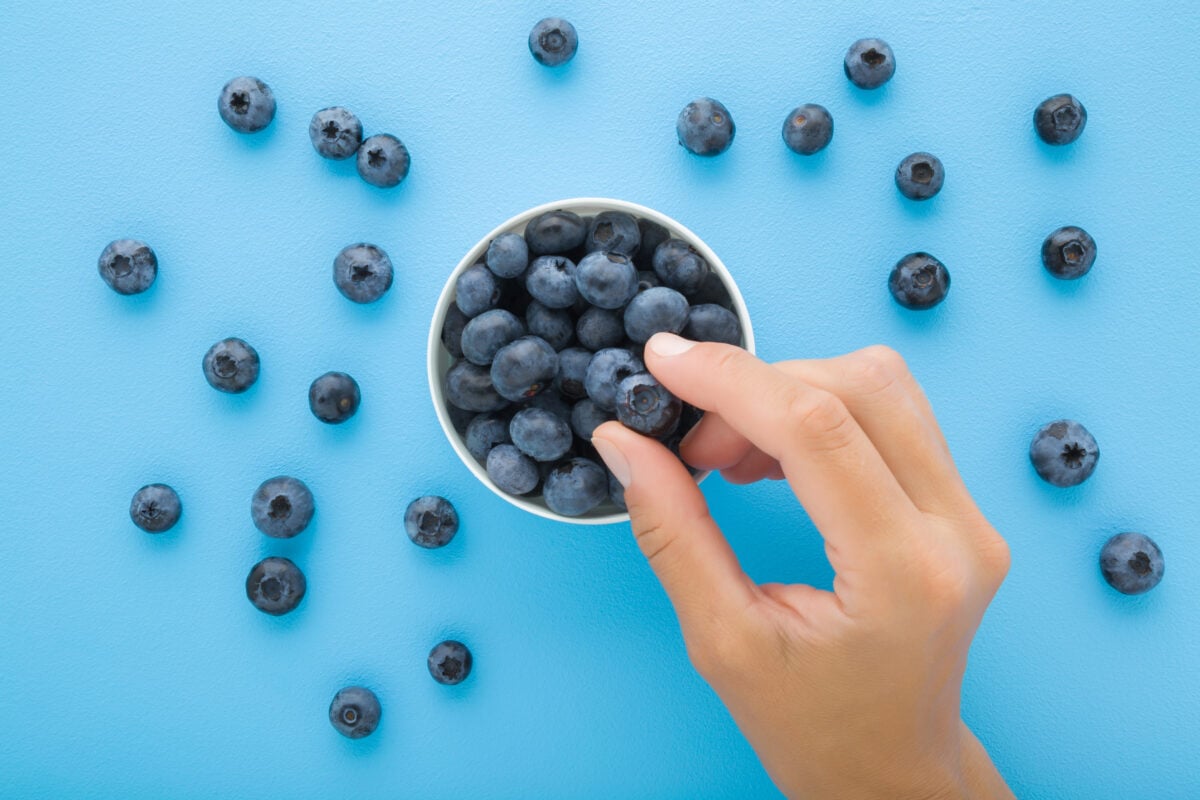 Photo shows someone's hand as they eat blueberries from a small bowl. The bowl is on a pale blue background scattered with blueberries.