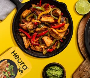 Photo shows a chopped up chicken fillet from Heura and Swap in a cast iron pan along with peppers and onions, against a yellow background, and alongside the product's new packaging