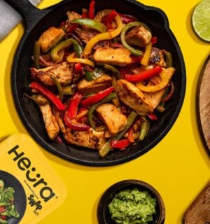 Photo shows a chopped up chicken fillet from Heura and Swap in a cast iron pan along with peppers and onions, against a yellow background, and alongside the product's new packaging