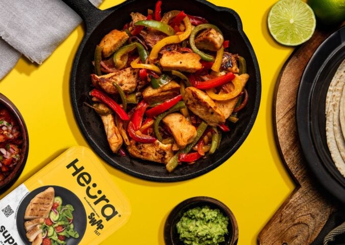 Photo shows a chopped up chicken fillet from Heura and Swap in a cast iron pan along with peppers and onions, against a yellow background, and alongside the product's new packaging