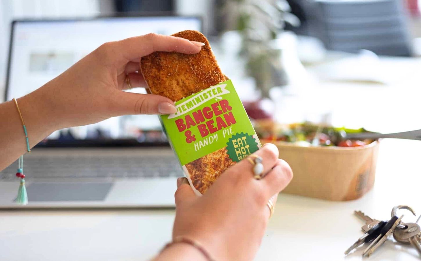 A person sat at a desk holding a vegan "banger & bean" handy pie by Piminister