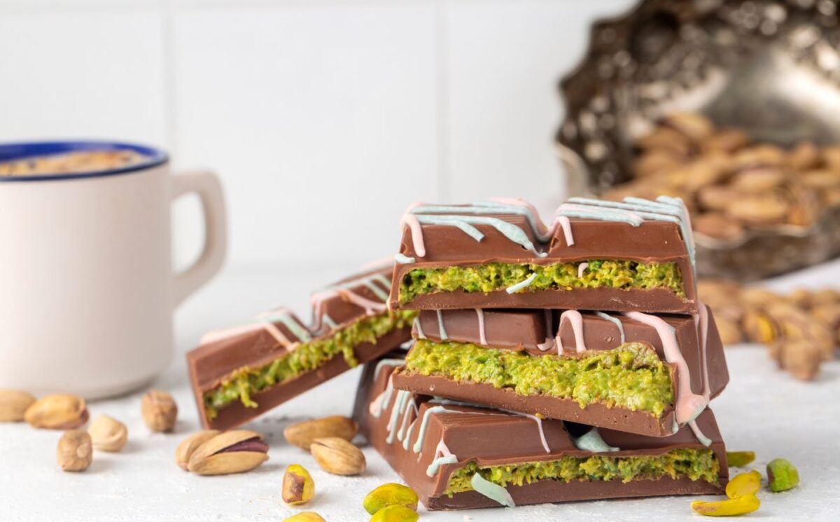 Photo shows a stack of Dubai Chocolate bar pieces, including a decorative pattern on the chocolate shell and a distinctive green filling, next to a cup of tea and pistachio shells