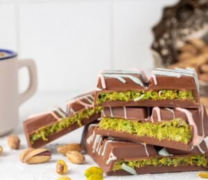 Photo shows a stack of Dubai Chocolate bar pieces, including a decorative pattern on the chocolate shell and a distinctive green filling, next to a cup of tea and pistachio shells