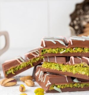 Photo shows a stack of Dubai Chocolate bar pieces, including a decorative pattern on the chocolate shell and a distinctive green filling, next to a cup of tea and pistachio shells