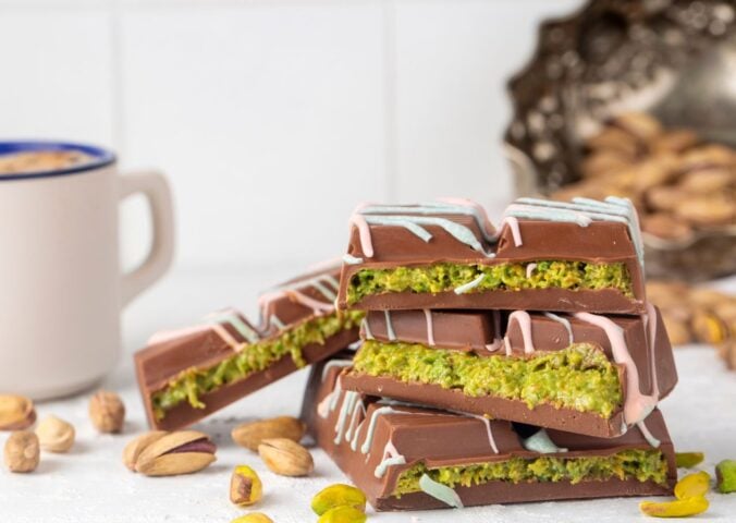 Photo shows a stack of Dubai Chocolate bar pieces, including a decorative pattern on the chocolate shell and a distinctive green filling, next to a cup of tea and pistachio shells